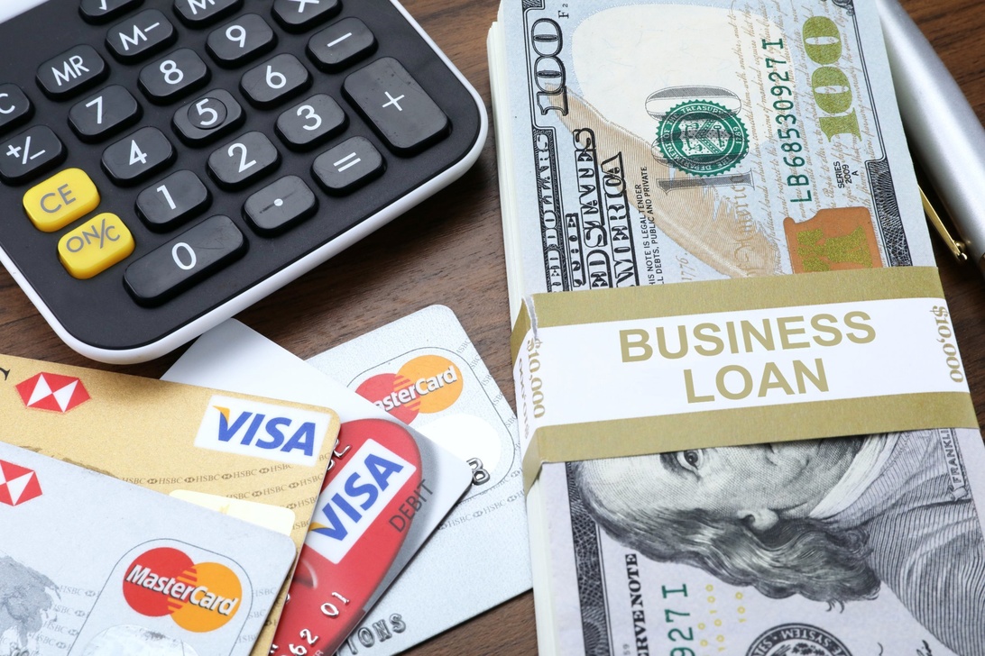 En bild som visar olika finansieringsalternativ för företag, inklusive en bunt sedlar märkt 'BUSINESS LOAN', en miniräknare och kreditkort.